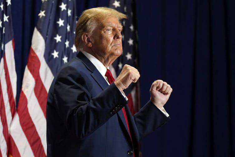 Donald Trump, debout, les poings serrés, avec un drapeau américain en arrière-plan