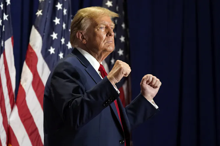 Donald Trump, debout, les poings serrés, avec un drapeau américain en arrière-plan