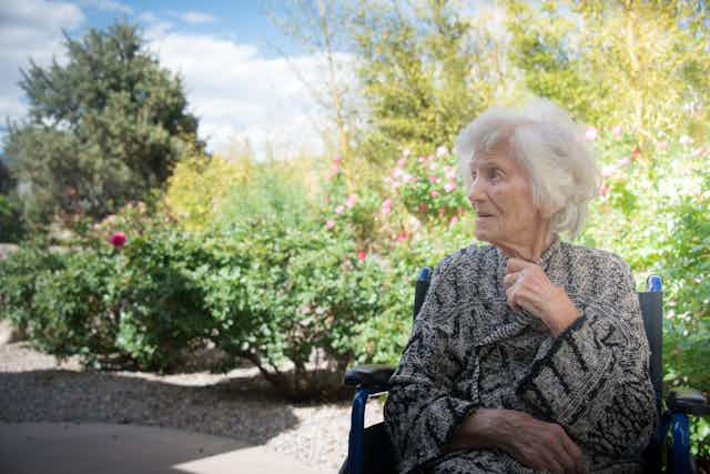 A senior woman in a wheelchair sits outside in a garden.