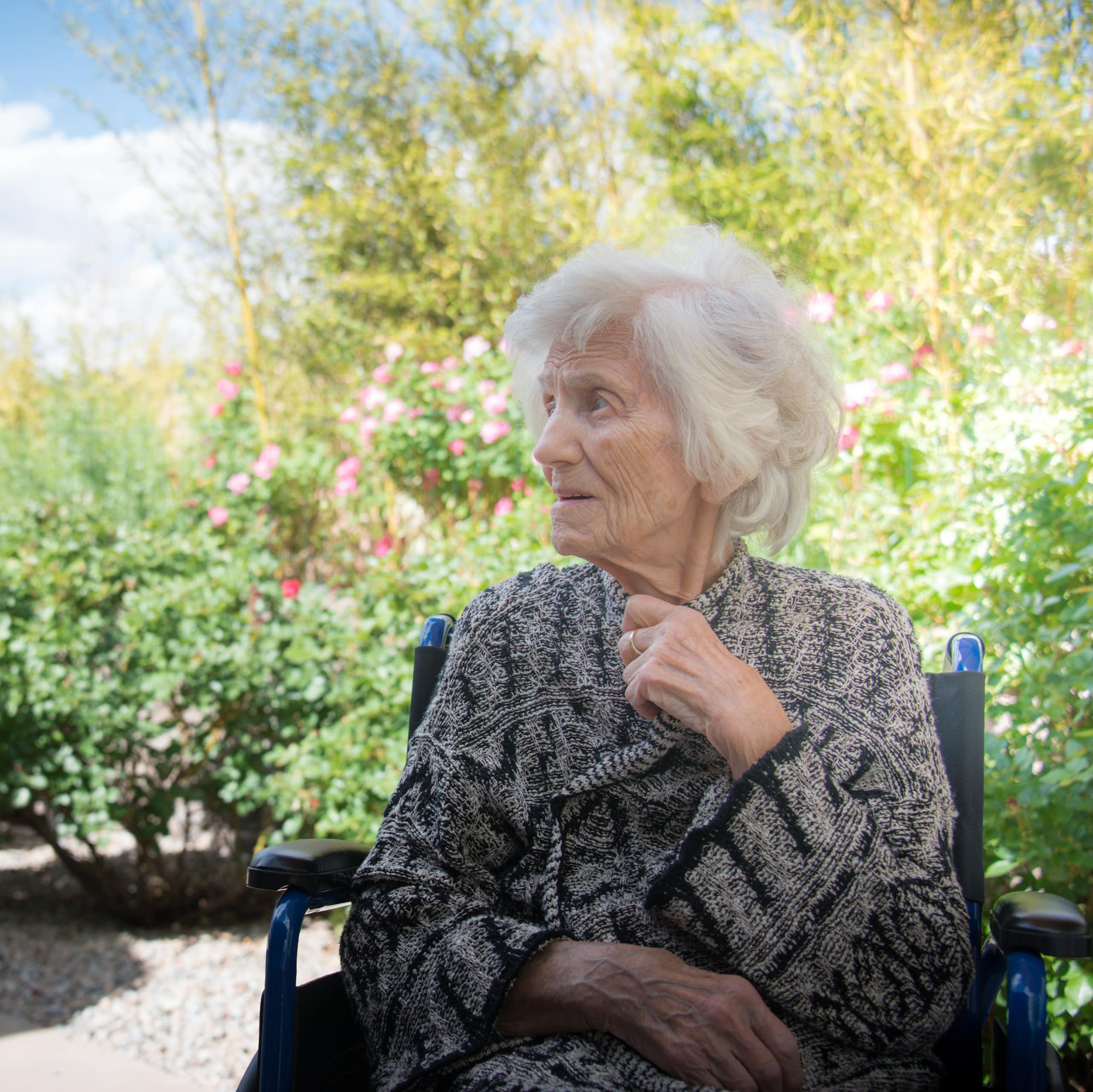 A senior woman in a wheelchair sits outside in a garden.