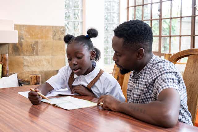A man seated at a table with a girl, both of them looking at an open book as the girl reads aloud