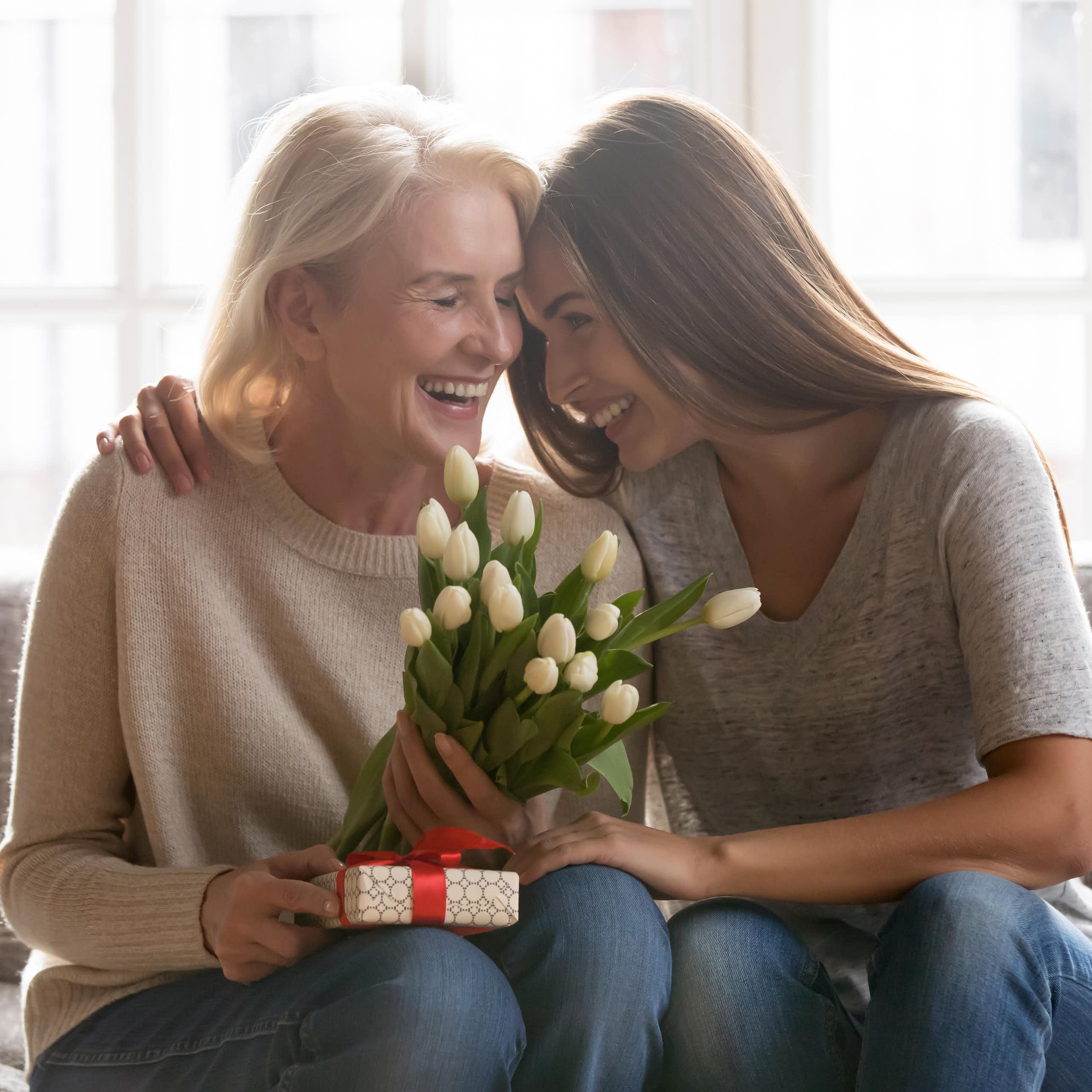 An older woman and her adult daughter smile and embrace on a sofa, the mother is holding a bouquet of white tulips and a wrapped gift