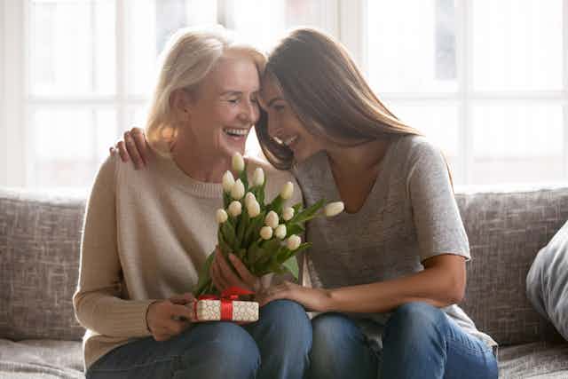 An older woman and her adult daughter smile and embrace on a sofa, the mother is holding a bouquet of white tulips and a wrapped gift
