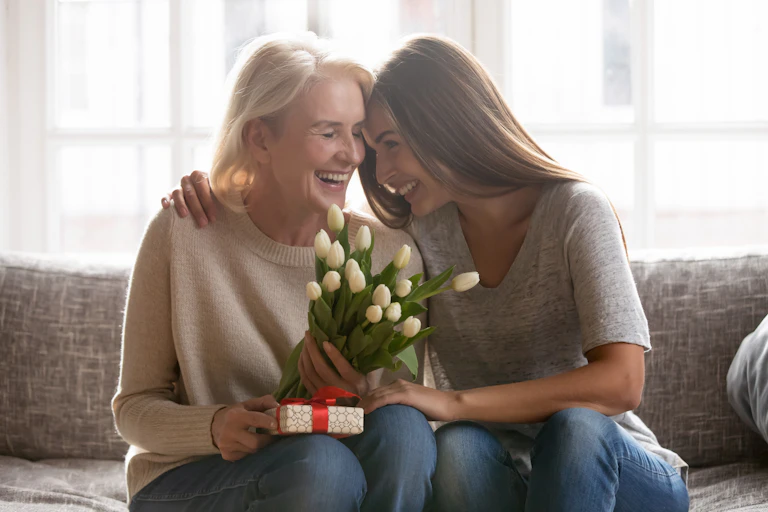An older woman and her adult daughter smile and embrace on a sofa, the mother is holding a bouquet of white tulips and a wrapped gift