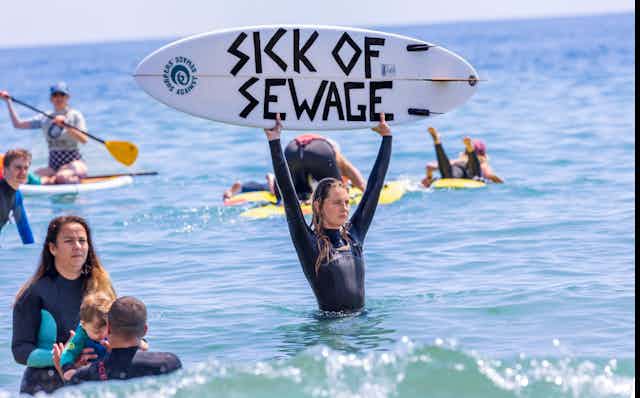 A female surfer in the sea holds up a surfboard with the words 'sick of sewage' written on it. 