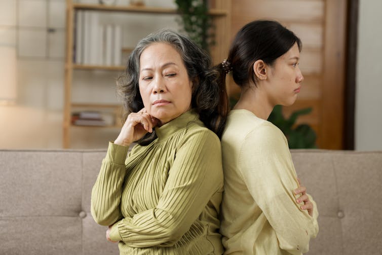 An older, Asian woman and her adult daughter stand back to back with arms crossed and unhappy expressions on their faces