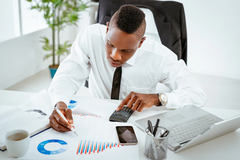 A young man in a white shirt with a black tie pores over graphs with a pen in one hand and a calculator in the other