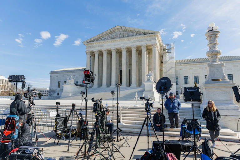 People set up cameras and microphones outside a stately building fronted with pillars.