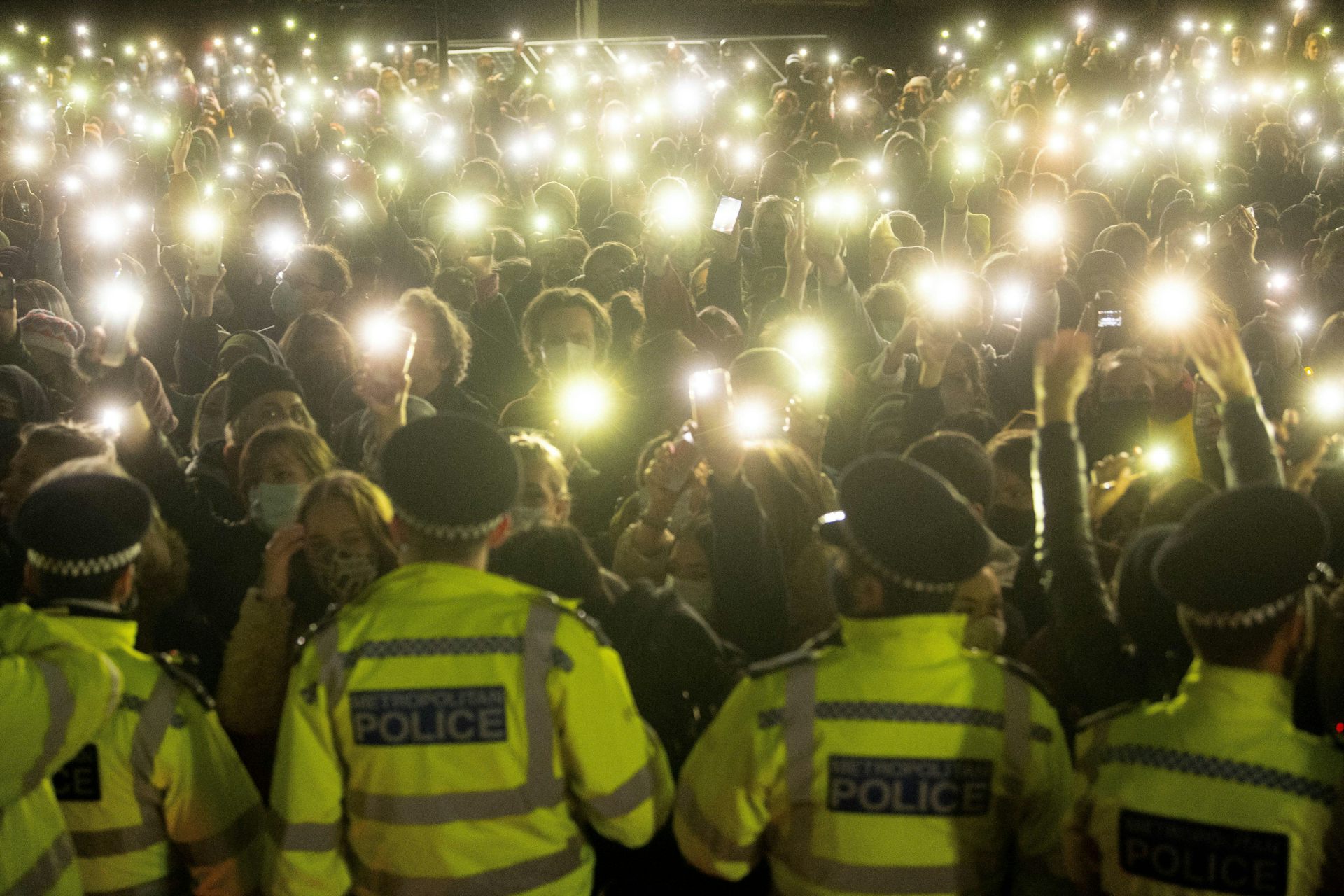 A row of Met police, seen from behind, face a crowd of people with their phone torches lit up at a vigil