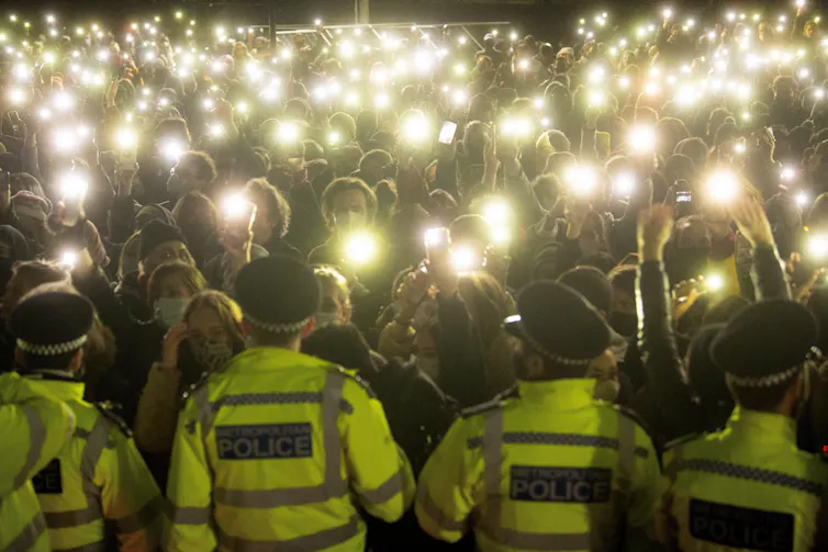 A row of Met police, seen from behind, face a crowd of people with their phone torches lit up at a vigil