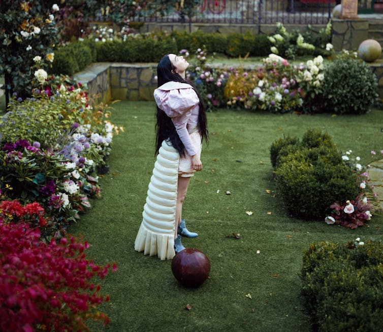 A woman in bronze ornate dress stares into the camera and reflection is in a mirror beside her.