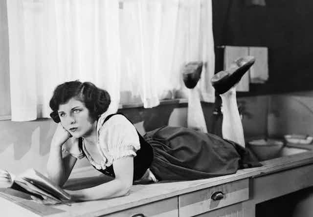 A woman lying on a kitchen sideboard reading a book, looking fed up.