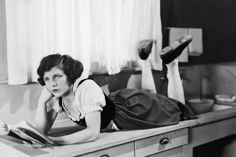 A woman lying on a kitchen sideboard reading a book, looking fed up.
