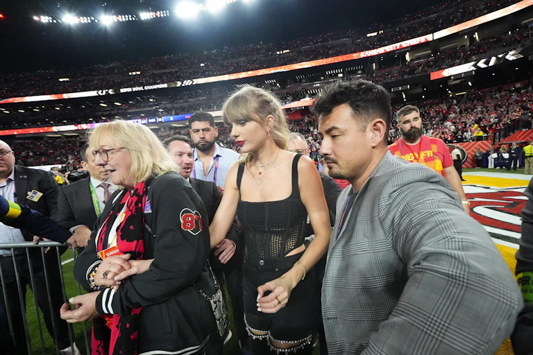 A blonde woman wears a black tank top and holds the hands of an older blonde woman next to her. She is flanked by a man on her other side and walks in a football field.