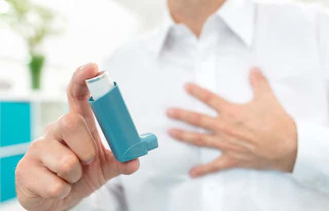 A man is seen from the neck down, grasping his chest with one hand and holding an asthma inhaler with the other.