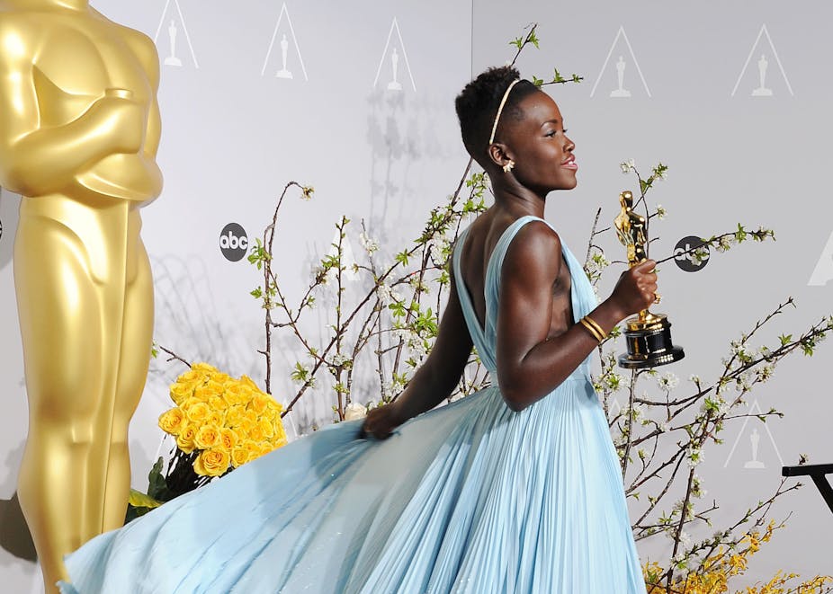 Young Black woman with short hair holds gold statuette while wearing a flowing powder blue dress.