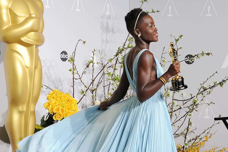 Young Black woman with short hair holds gold statuette while wearing a flowing powder blue dress.