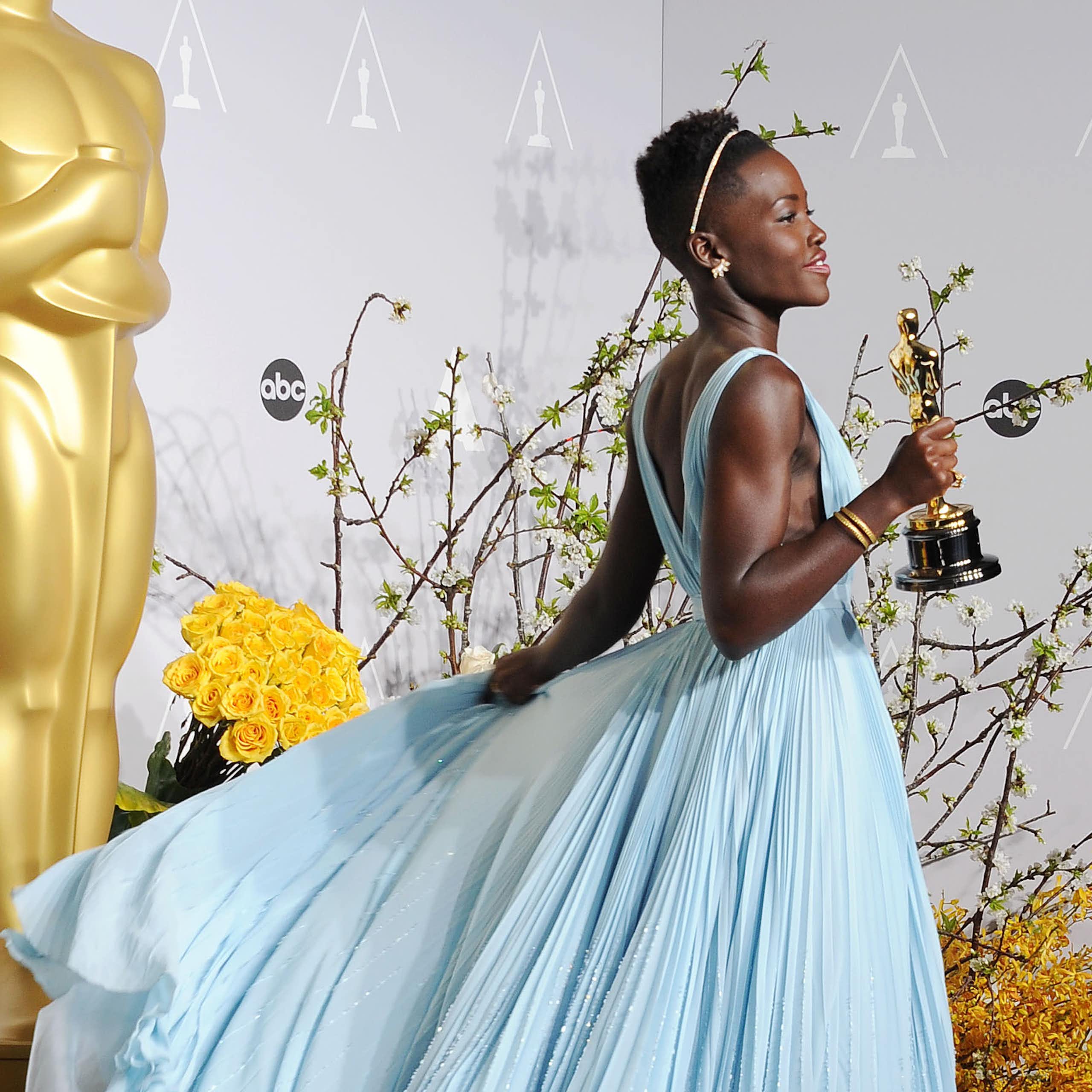 Young Black woman with short hair holds gold statuette while wearing a flowing powder blue dress.