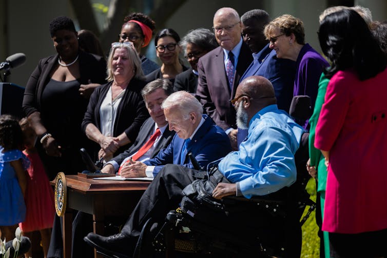 President Biden sitting at a desk signing with a crowd gathered around him