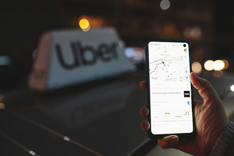 Uber app displayed on smartphone held in a hand in front of taxi with an Uber sign on top of a car