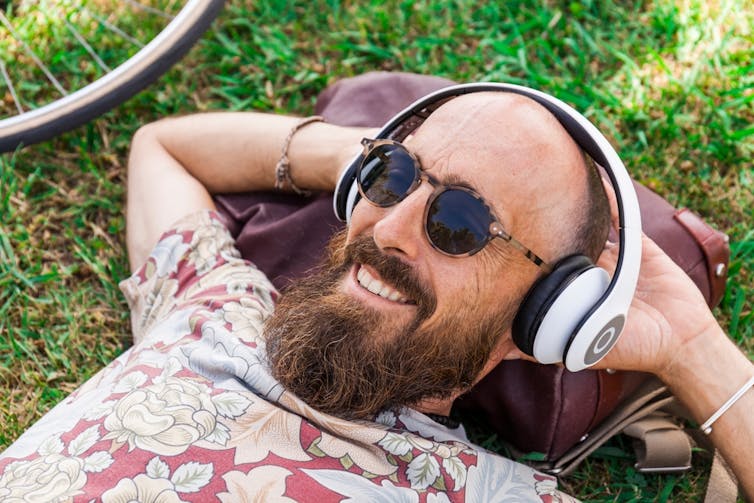 A happy and handsome man with receding hairline relaxes in a park while listening to headphones.