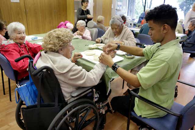 Elderly women, including one in a wheelchair, do art projects at a nursing home; a younger man in the foreground helps.