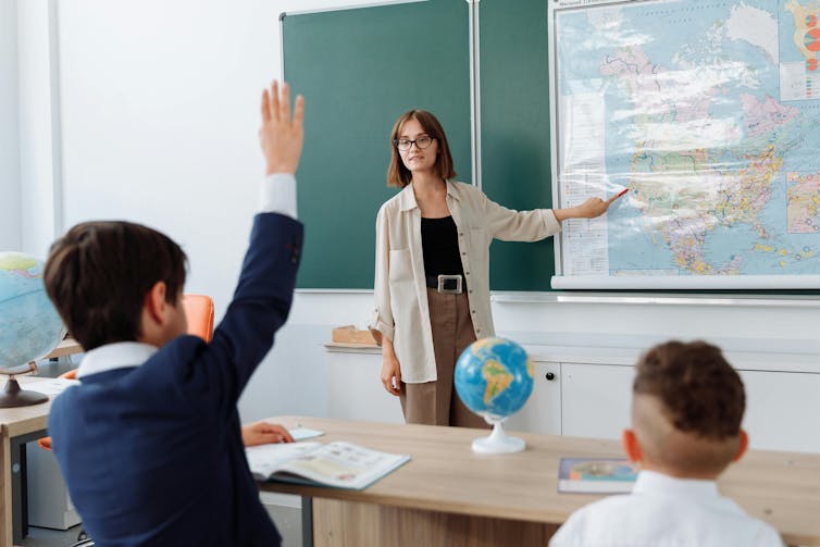 A boy in a school uniform raises his hard. A female teacher points to a map on a board.