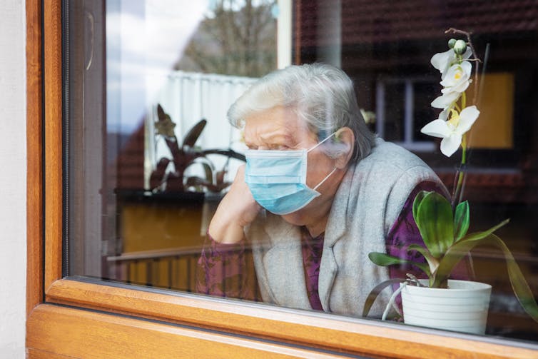 A lonely older woman looking out a window while wearing a medical mask.