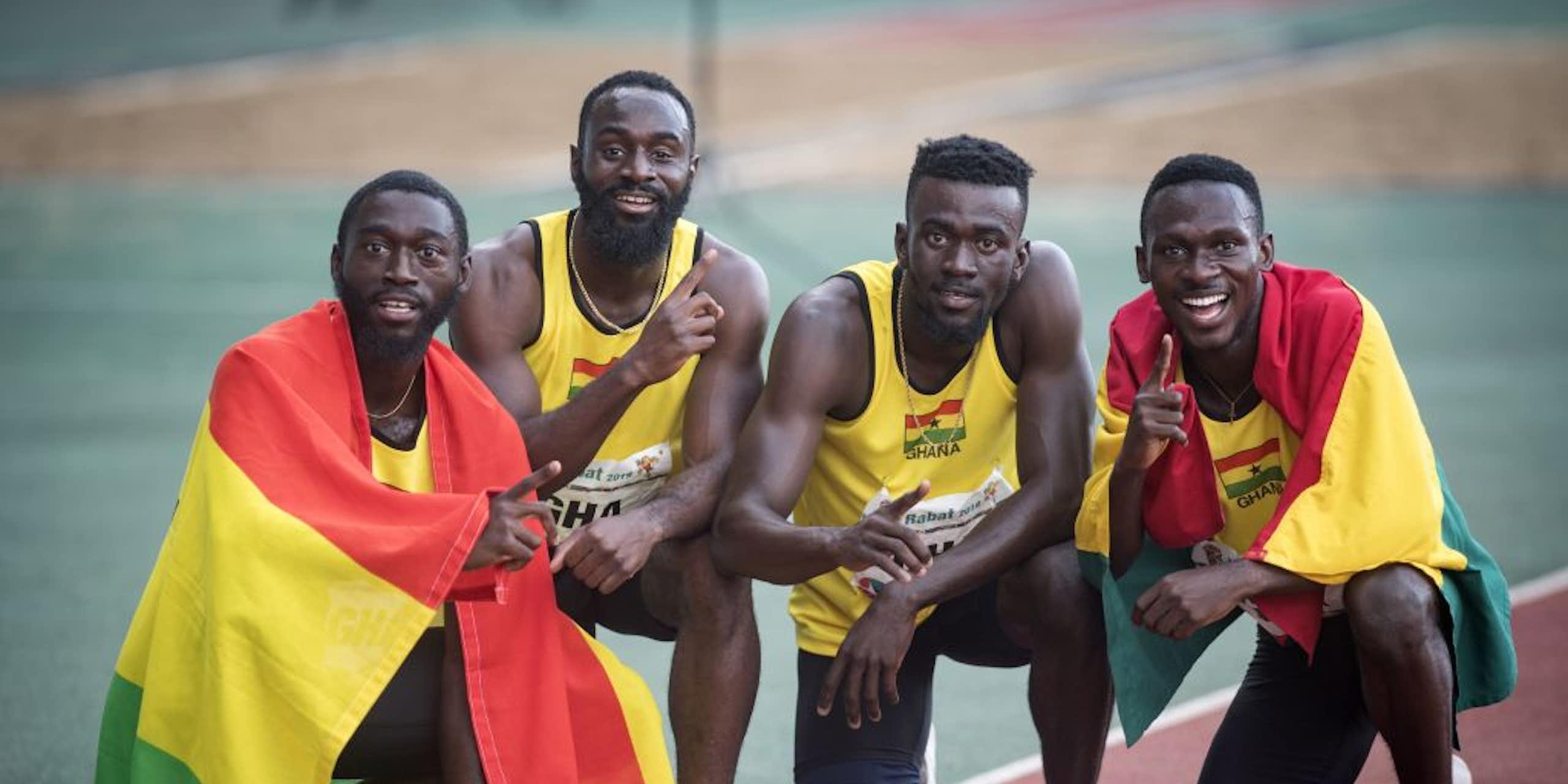Four men in athletics gear kneel and crouch on a running track, two of them draped in the green, red and yellow flags of Ghana.