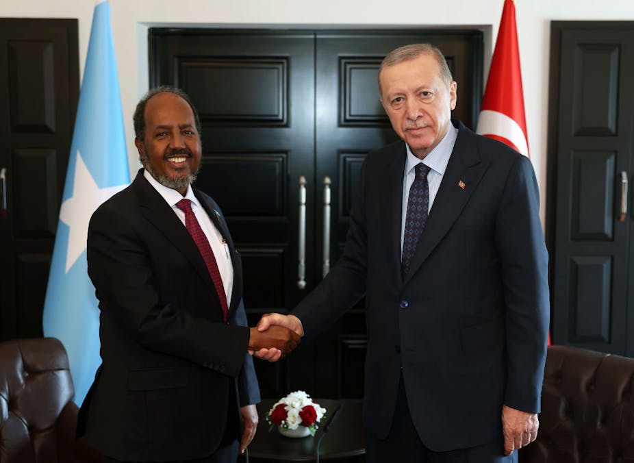 Two men shaking hands as they look at the camera with blue and red flags on either side of them