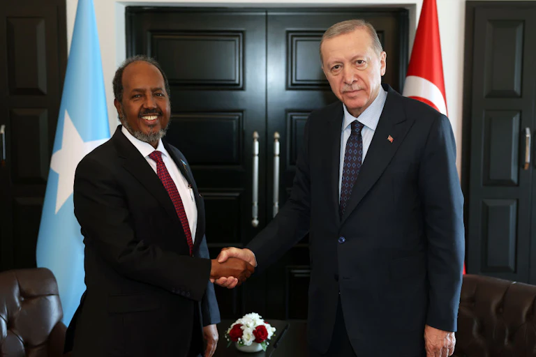 Two men shaking hands as they look at the camera with blue and red flags on either side of them