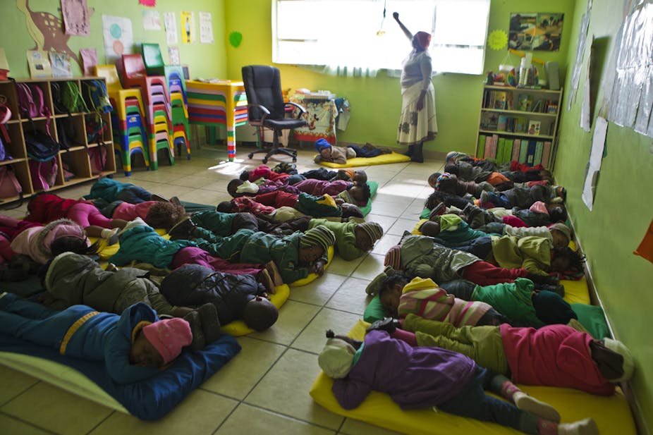 A woman in a colourful classroom pulls down a blind while children settle down on mattresses