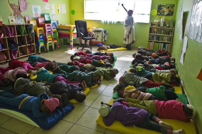 A woman in a colourful classroom pulls down a blind while children settle down on mattresses