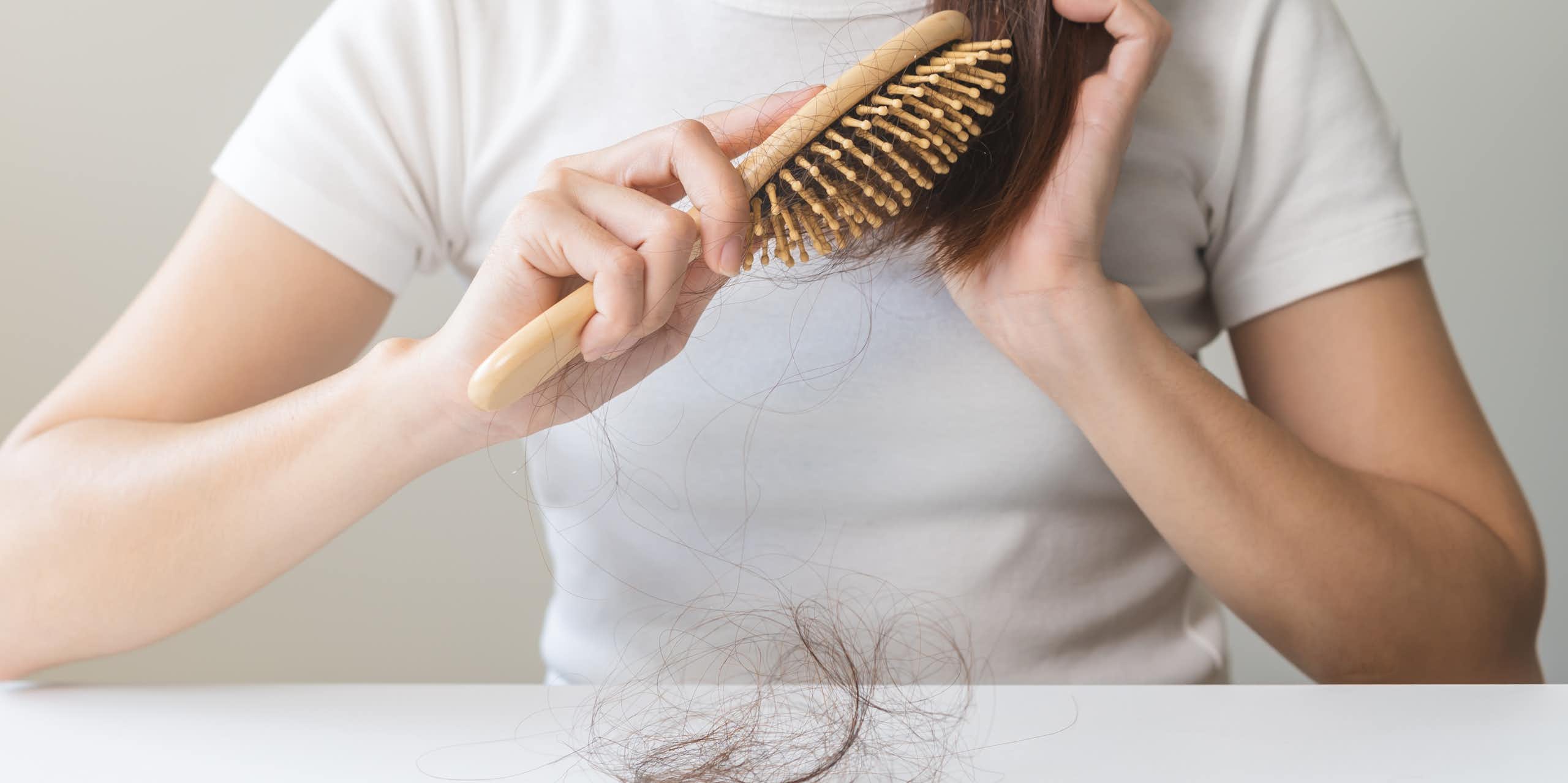 A woman brushes her hair using a wooden hair brush. There's a clump of hair on the table in front of her.