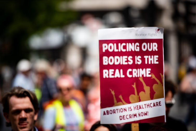 A protest sign reads 'policing our bodies is the real crime, abortion is healthcare, decrim now'