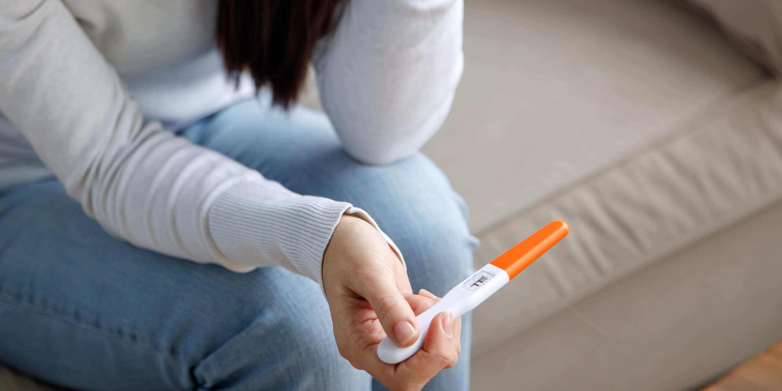 A woman sitting on a couch holding a pregnancy test, her face isn't visible but her body language suggests she is not happy