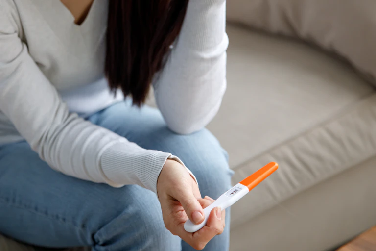 A woman sitting on a couch holding a pregnancy test, her face isn't visible but her body language suggests she is not happy