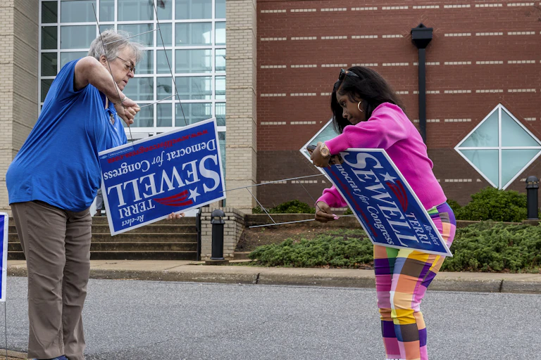 Two people work to set up campaign signs, some sideways and others upside down.