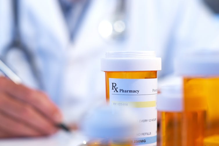 A person in a white coat out of focus in the background with a prescription bottle in the foreground