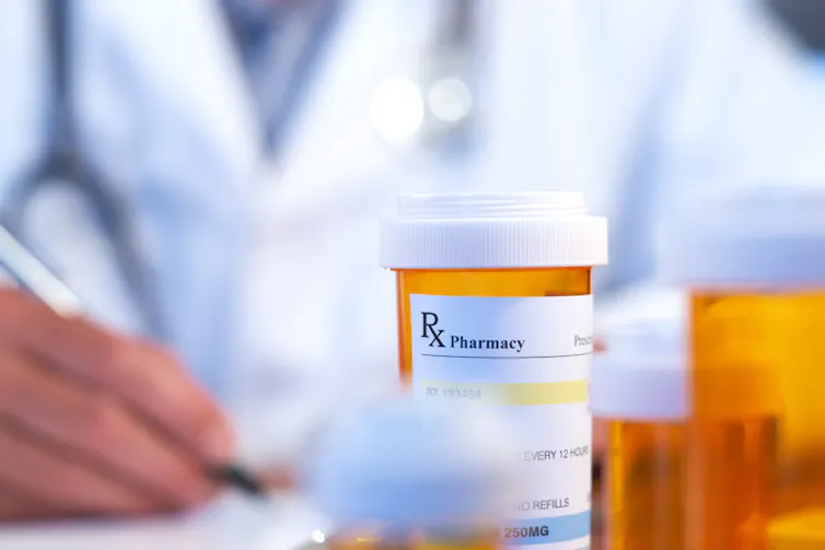 A person in a white coat out of focus in the background with a prescription bottle in the foreground
