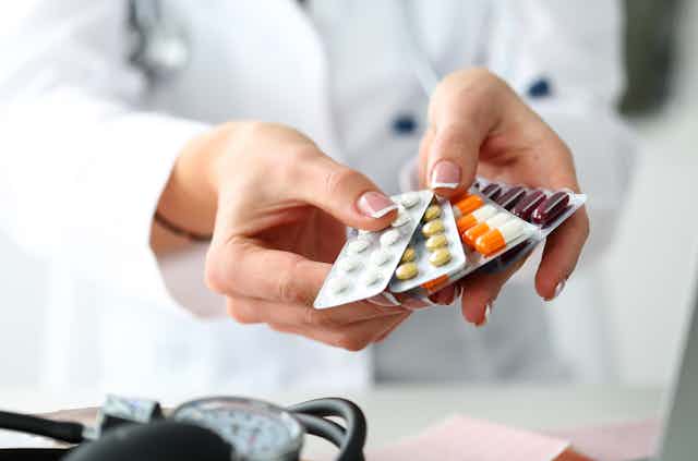 A person in a white coat out of frame holding out a selection of blister-packed drugs