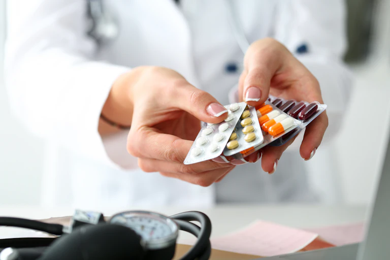 A person in a white coat out of frame holding out a selection of blister-packed drugs