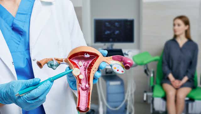 Mão de médico aponta modelo do aparelho reprodutivo feminino com mulher ao fundo em consultório