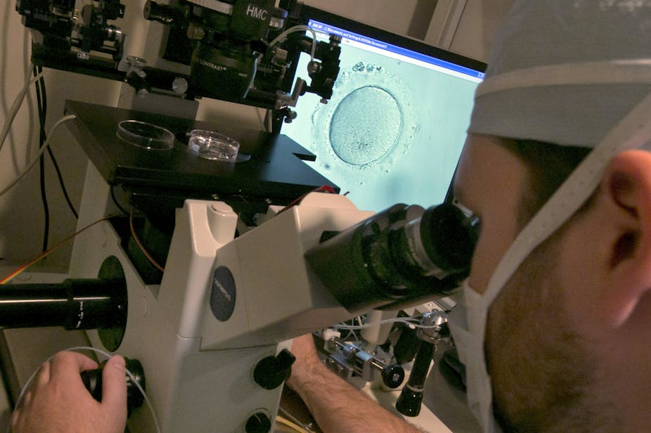 A person in a medical mask and cap looks through a microscope, as the screen shows a gray-blue dot.