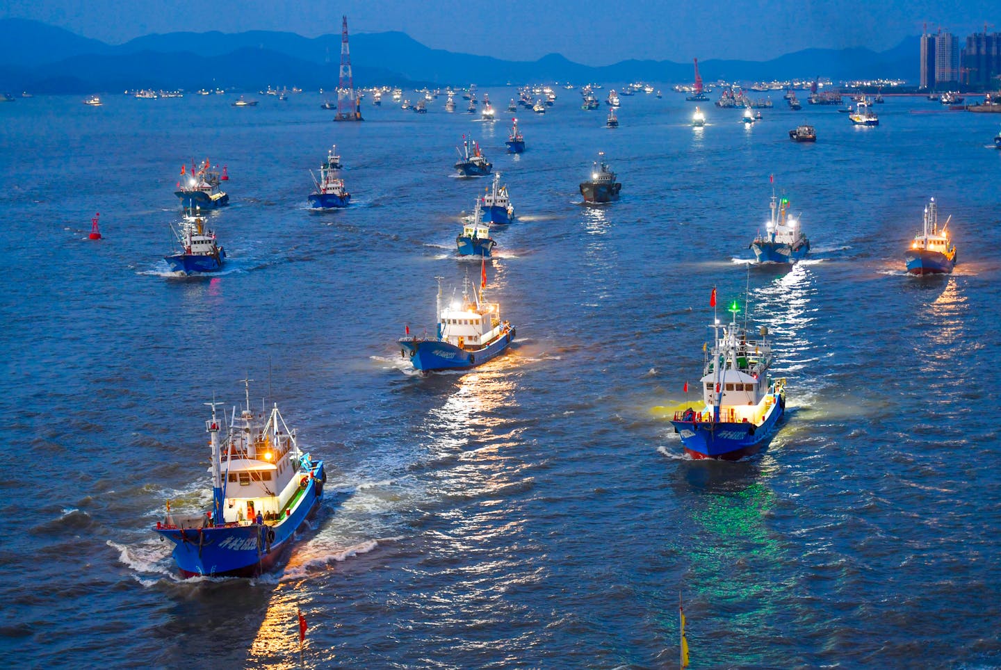 Dozens of fishing boats move out of an urban harbor