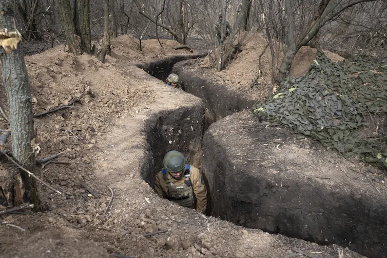 Soldiers wearing helmets move down a narrow trench in a woodland area