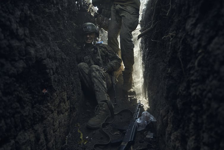 A soldier sits in a narrow trench as another walks past