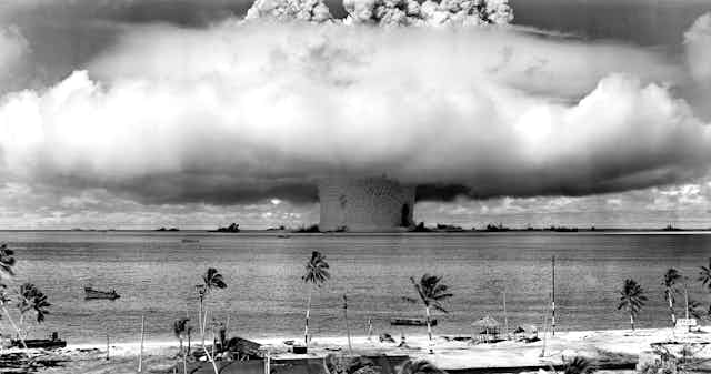 Black and white photo of a mushroom cloud in the ocean, as seen from the shore.