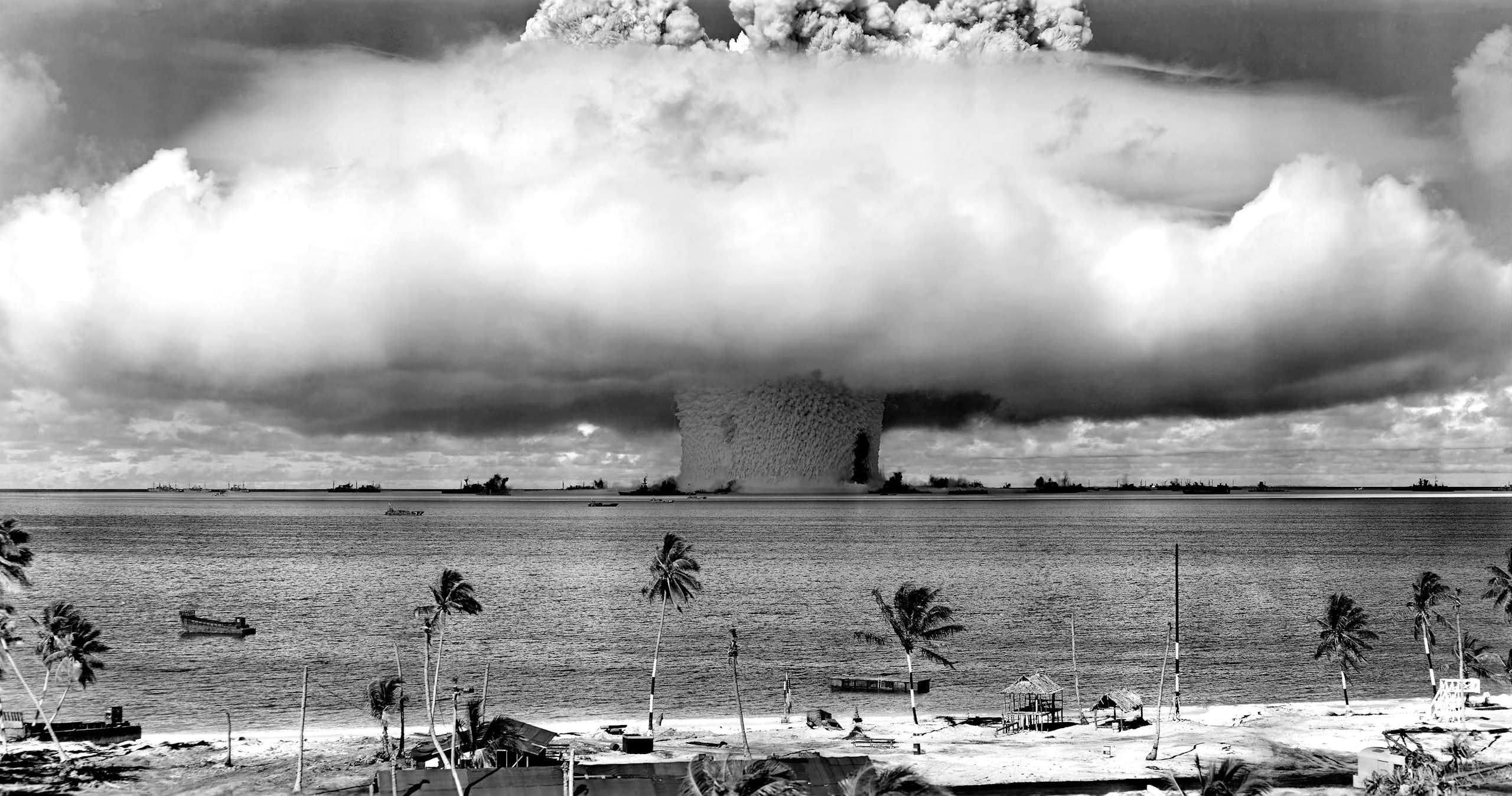 Black and white photo of a mushroom cloud in the ocean, as seen from the shore.