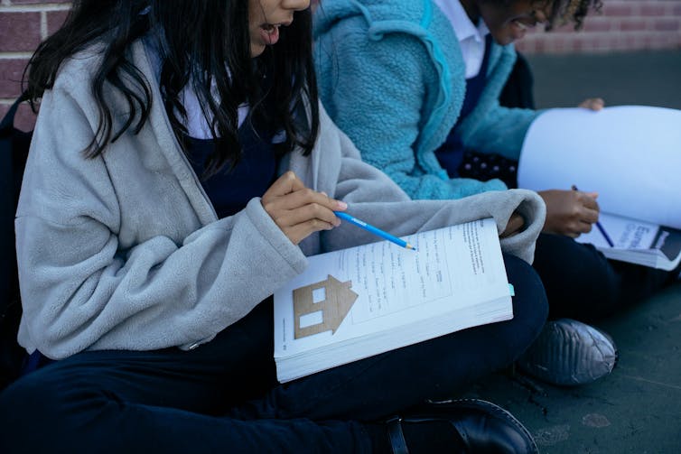 Two young students sit cross legged in a playground, looking at work books.